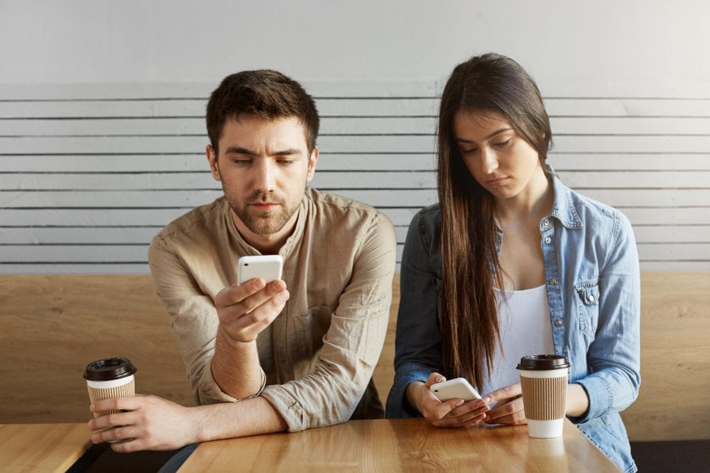 Married couple distracted on their phones during dinner, symbolizing emotional drift caused by normalized habits