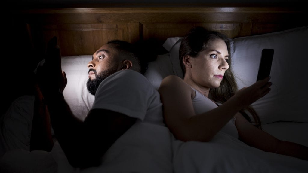 Married couple disengaged in bed, choosing screen time over intimacy.